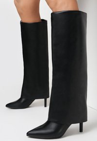 Bottes noires montantes en cuir lisse. Dotées d'un bout pointu et d'un talon aiguille. Le tige des bottes a un design épuré et minimal.