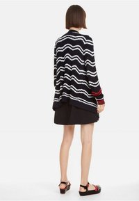 Cardigan a righe blu navy e bianche con un motivo ondulato, caratterizzato da una vestibilità ampia, maglia testurizzata, accenti rossi sulle maniche, abbinato a una gonna nera.