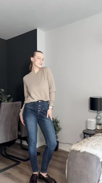 Top bege de manga comprida com corte descontraído, jeans skinny azuis com fecho frontal de botões e sapatos pretos brilhantes. Ambiente interior com tons neutros.