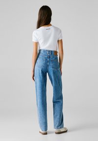 Mujer vestida con una blusa blanca de manga corta y jeans de tiro alto de color azul claro, de pie con la espalda hacia la cámara frente a un fondo claro y liso.