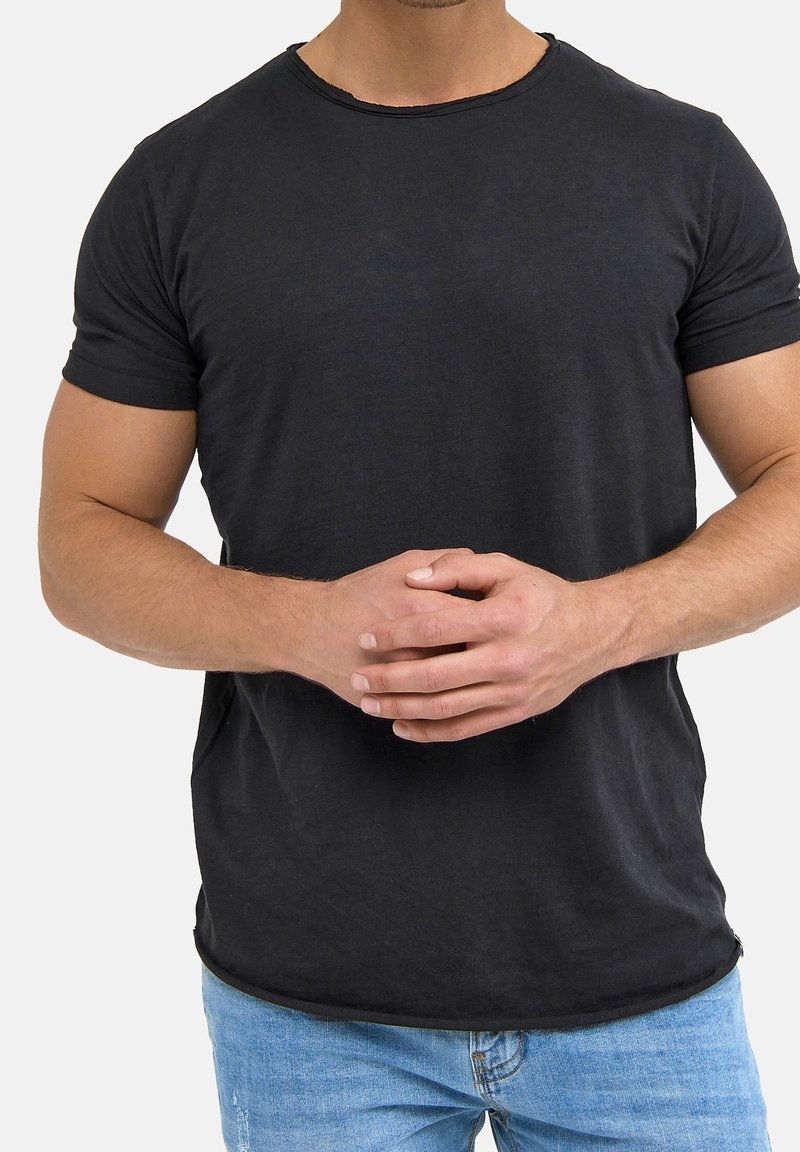 Homme portant un t-shirt noir à manches courtes et un jean bleu, debout les mains croisées à la taille sur un fond uni.
