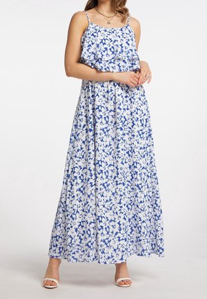 Donna che indossa un abito lungo senza maniche a fiori blu e bianco con spalline sottili e sandali bianchi con punta aperta, in piedi con le mani giunte.