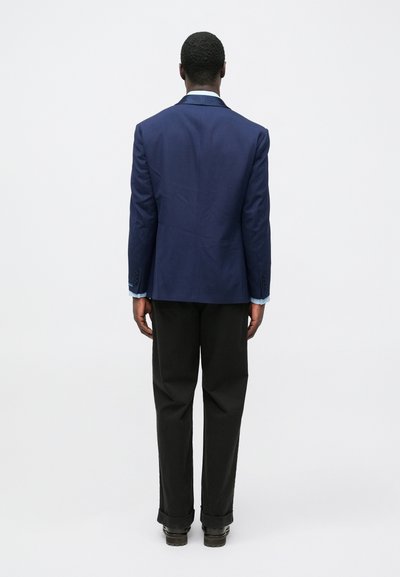 Modrý sako na mieru so zvýšenými ramenami, jedným rozparkom a manžetami na gombíky, v kombinácii s čiernymi nohavicami a pevnými čiernymi topánkami, pohľad zozadu.