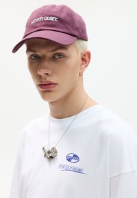 Casquette en coton bordeaux avec "NEVER QUIET" brodé en blanc ; t-shirt blanc avec un motif graphique bleu ; collier en pendentif argenté.