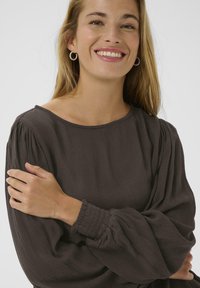Mørkebrun bluse med rund halsudskæring, lange puffærmer og smockede manchetter. Stoffet har et blødt, tekstureret udseende.