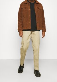 Chaqueta de shearling marrón con cierre de botones, camiseta negra debajo, pantalones beiges y botines negros. Pose de pie en plano sobre un fondo blanco.