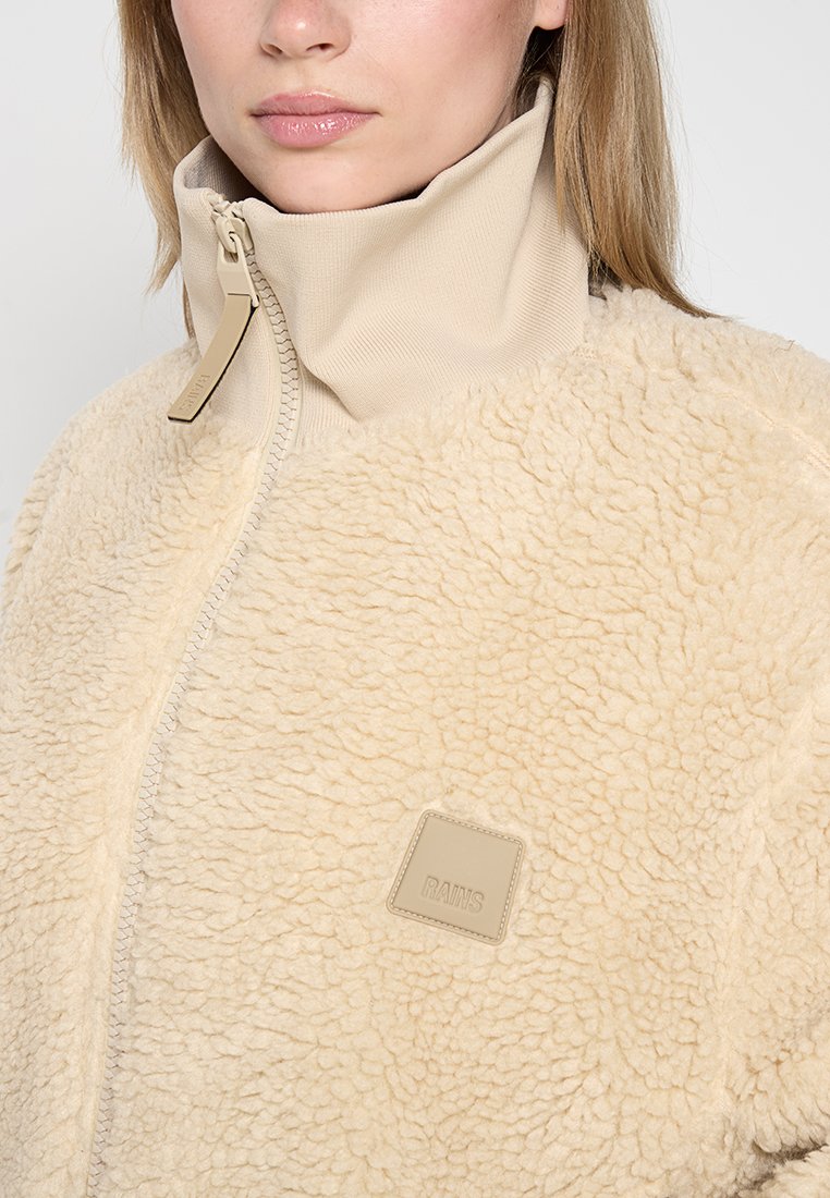 Chaqueta de forro polar beige con cuello alto, cierre de cremallera y superficie texturizada con un parche cuadrado de marca.