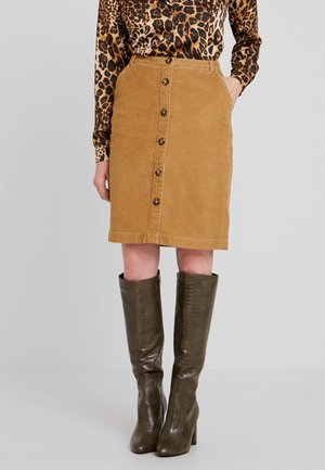 Kvinde iført en tan knap-front fløjlsnederdel, leopardprint bluse og mørkebrune, knæhøje teksturerede støvler, stående mod en ensfarvet baggrund.