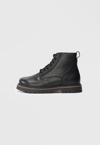 Botín de cuero negro con cordones, de superficie texturizada y suela rugged, visto desde el lado exterior sobre un fondo plano.
