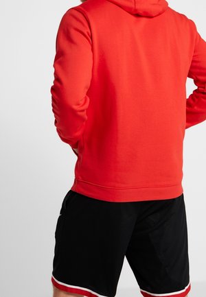 Mann trägt einen leuchtend roten Hoodie und schwarze Sportshorts mit weißem und rotem Rand, steht mit den Händen in die Hüften gestützt und schaut weg.