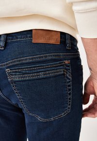 Dark blue denim jeans feature brown leather brand patch, contrast stitching, and two back pockets with curved seams.