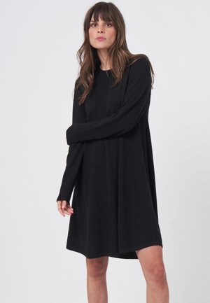 Junge Frau mit braunen Haaren, die ein lockeres schwarzes Langarmkleid trägt, steht vor einem einfachen hellen Hintergrund.