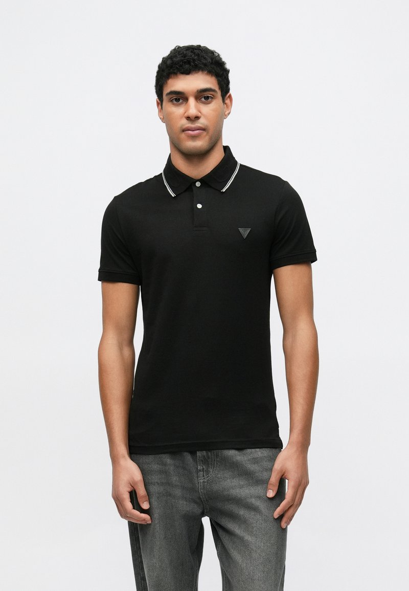 Μαύρο πόλο πουκάμισο με στενή γραμμή, διαθέτει αντίθετο λευκό γιακά, πατιλέτα με δύο κουμπιά και μικρό τριγωνικό λογότυπο στο στήθος.