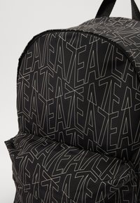 Zaino nero con un motivo testuale geometrico ripetuto in grigio chiaro. Presenta una parte superiore arrotondata e una tasca frontale con zip.