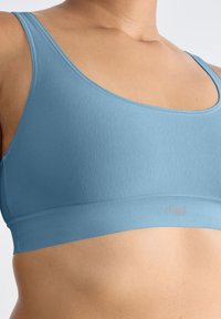 Sininen urheilu-toppi, jossa on saumaton muotoilu, leveät hihnat ja pehmeä joustava vyö alareunassa, jossa on pieni logo. Liukas pinta.