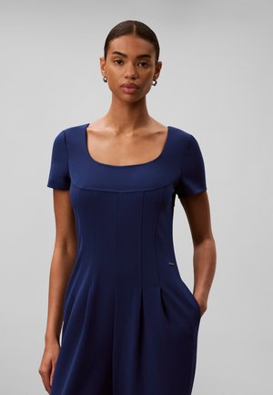 Jeune femme aux cheveux courts et foncés, portant une robe bleu marine ajustée avec des manches courtes et un décolleté carré, se tenant devant un fond uni.