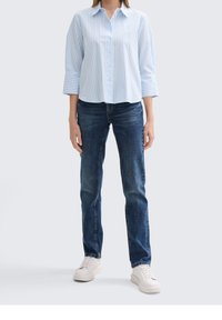 Camisa de botones a rayas en azul claro y blanco con puños anchos, combinada con jeans azul oscuro y zapatillas blancas. Ajuste casual, con un detalle mínimo.