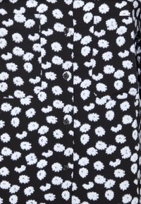 Camisa negra con patrones florales blancos, dos bolsillos en el pecho y botones negros. La tela parece suave con una textura ligera.