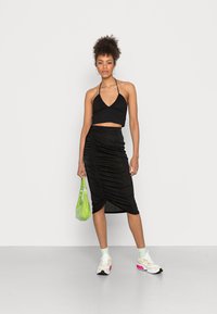 Top crop preto com alças halter combinado com uma saia midi preta drapeada. A modelo segura uma pequena mala verde. Ténis brancos com detalhes em rosa.