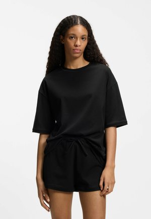 Woman with long curly hair wearing a loose black T-shirt and matching black shorts, standing against a plain light background.