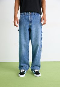 Pantalons cargo en denim bleu clair avec une coupe ample, dotés de poches latérales, de détails cousus et d'une fermeture à bouton. Portés avec des sneakers noires.