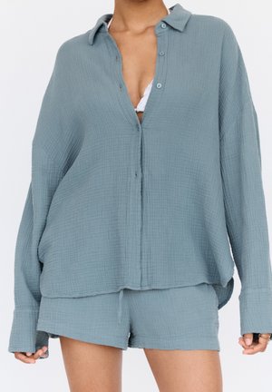 Chemise bleue à boutons ample avec texture et shorts assortis portés sur un bralette blanc par une personne debout.