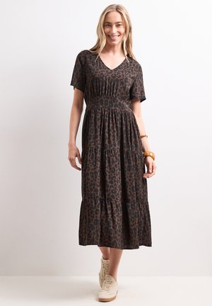 Femme souriante portant une robe midi imprimé léopard marron et noir à manches courtes et baskets beige sur un fond clair uni.