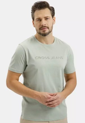 Mężczyzna o krótkich ciemnych włosach i brodzie, ubrany w jasno szarą koszulkę "Cross Jeans", stoi z luźno splecionymi rękami z przodu.