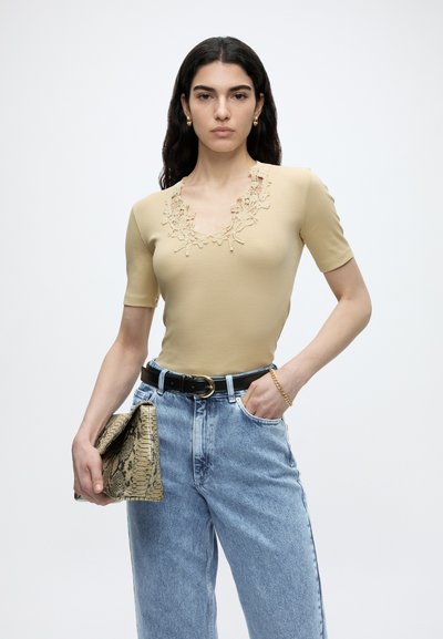 Γυναίκα με μακριά σκουρόχρωμα μαλλιά που φοράει μπεζ τοπ με δαντέλα και ανοιχτό μπλε τζιν, κρατά ένα τσαντάκι από φίδι και φοράει χρυσό βραχιόλι και σκουλαρίκια.