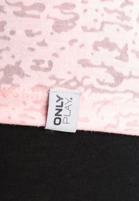 Jasnoróżowa tkanina z subtelnym wzorem kamuflażu, z czarnym dolnym obszyciem i białą metką z napisem "ONLY PLAY".