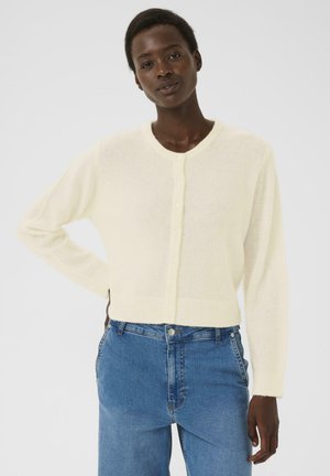 Femme aux cheveux naturels courts portant un cardigan blanc à boutons et un jean taille haute bleu, se tenant avec une main sur la hanche.