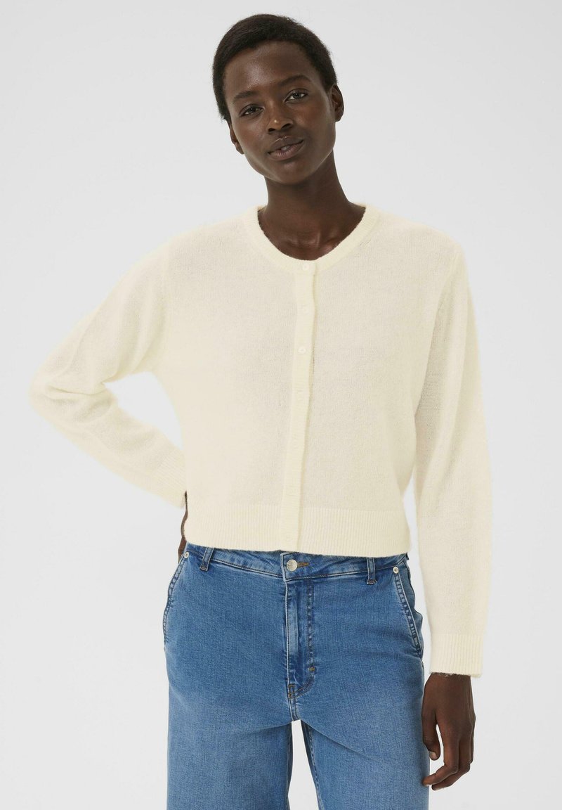 Femme aux cheveux naturels courts portant un cardigan blanc à boutons et un jean taille haute bleu, se tenant avec une main sur la hanche.