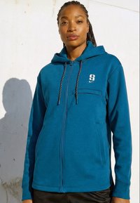 Sieviete, kas valkā tirkīzzilu džemperi ar rāvējslēdzēju, kapuci un melnām un baltām auklām, stāv pret gaišu sienu dabīgā saules gaismā.