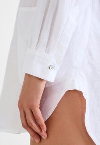 Hvit lin skjorte med lange ermer, kneppede mansjetter, lett gjennomsiktighet og et design med høy-lav nederkant. Har en sideslitt for ekstra detaljer.