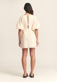 Crème kleurige jurk met puff mouwen, gaatjespatroon en een sleutelgat achterzak die met een strik is vastgemaakt. Tot boven de knie, getailleerd, eenvoudig ontwerp.