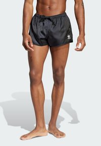 Schwarze Badehosen aus einem glänzenden Material, mit einem elastischen Taillenbund mit Kordelzug und einem kleinen weißen Logo an der Seite.