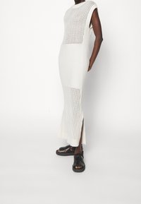 Gebreide witte jurk met geribbelde textuur en geperforeerde details; heeft een rechte silhouet en zijsplitten, gecombineerd met zwarte sandalen.