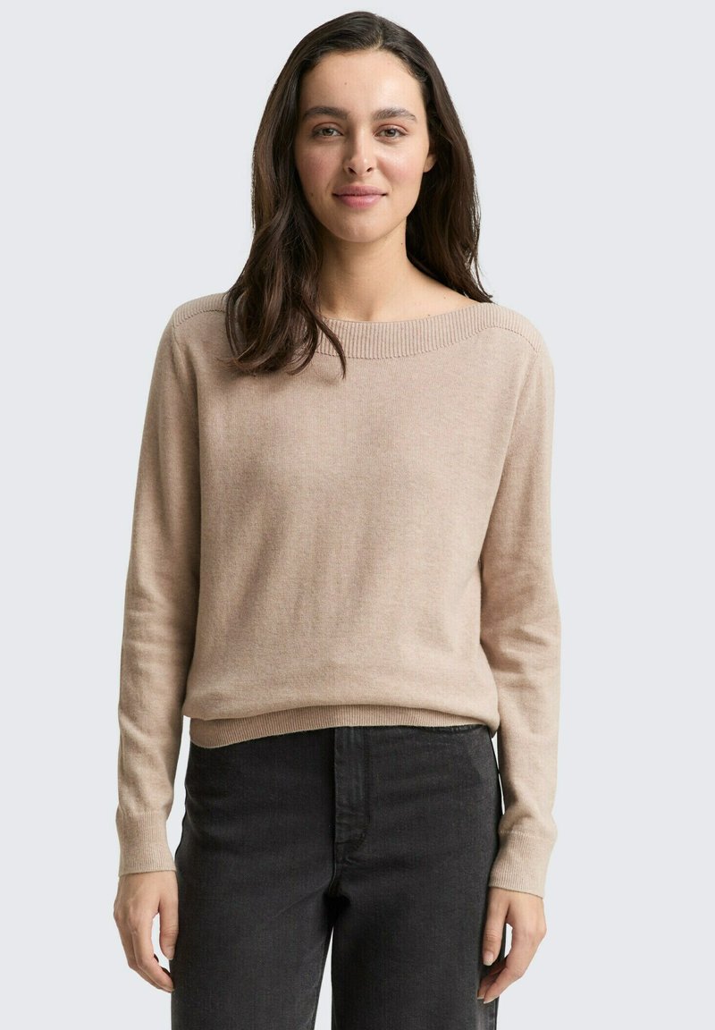 Vrouw met lang donker haar, gekleed in een beige trui en zwarte jeans, staand tegen een effen lichtgrijze achtergrond.