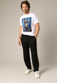 Man met krullend haar, die een wit T-shirt draagt met een grafiek van een Doberman hond in een zonnebril, zwarte broek en witte sneakers, staand met handen in zijn zakken.