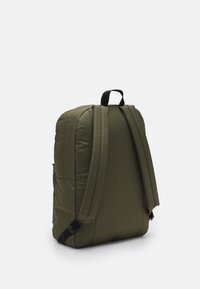 Mochila acolchada verde con correas negras ajustables, forma rectangular, bolsillo frontal y asa superior. Textura suave, con detalles de superficie acolchada.