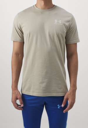 Mężczyzna w beżowej koszulce Under Armour i niebieskich spodenkach Under Armour stoi na jednolitym tle.
