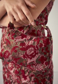 Hand hält eine kleine Handtasche mit roter Blumenstickerei, passend zu einem Kleid mit ähnlichem Muster.