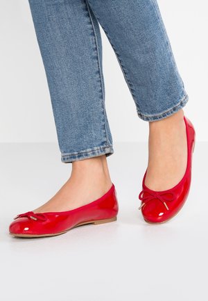 Pieds portant des ballerines rouges brillantes avec des nœuds, associées à un jean bleu denim sur un fond clair uni.