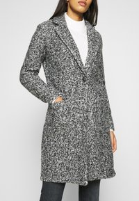 Grå och svart texturerad lång kappa med slagkrage, enkel knappstängning och sidofickor, lager-p å-lager över en vit tröja.