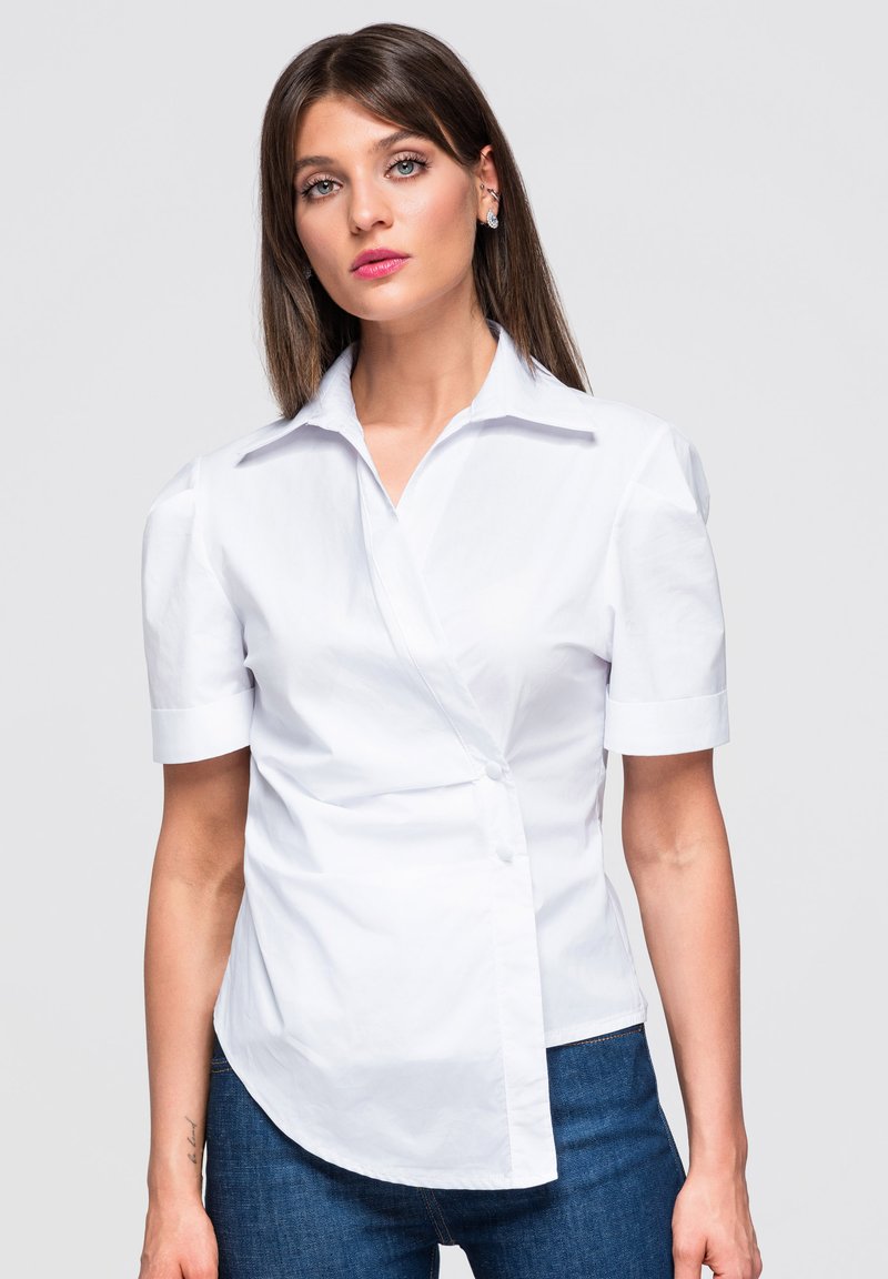 Femme aux cheveux bruns et raides porte un chemisier blanc à manches courtes avec un design asymétrique des boutons et un jean bleu sur un fond uni.