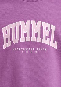 Lila sweatshirt med vit text "HUMMEL" och "SPORTSWEAR SINCE 1923." Materialet är mjukt med ribbad halsringning och slät yta.