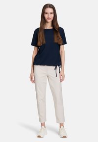 Granatowy top z krótkim rękawem o kwadratowym dekolcie i sznurkiem w dolnej części, zestawiony z beżowymi spodniami o zwężanej nogawce i białymi sneakersami.