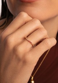 Goldring mit einem wellenförmigen Band, das eine Reihe von bunten Edelsteinen, darunter rosa und rot, auf einem strukturierten Hautton zeigt.