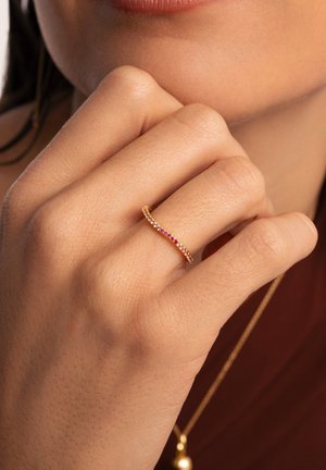 Bague en or avec un anneau ondulé, présentant une rangée de pierres précieuses colorées incluant des teintes roses et rouges, sur un fond de teinte de peau texturée.