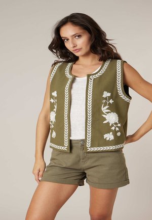 Femme portant un gilet sans manches brodé vert olive, un débardeur blanc, et un short assorti vert olive roulé, posant avec la main sur la hanche.